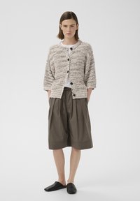 Femme portant un cardigan beige en maille avec des boutons noirs, un haut blanc, un short ample olive au genou et des chaussures noires sans lacets, debout les mains dans les poches.
