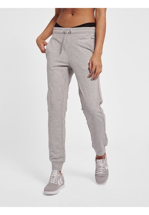 Personne portant un pantalon de survêtement gris avec une taille à cordon et des chevilles élastiquées, associé à des baskets grises et blanches sur fond blanc.