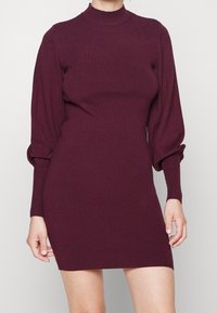 Abito a maglia a coste in Borgogna, con collo alto, vestibilità aderente e maniche lunghe a sbuffo. Il tessuto ha una texture liscia con polsini elastici.