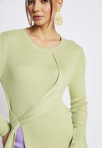Maglione a coste verde chiaro con design avvolgente, maniche lunghe e un pannello laterale con un pezzo di tessuto viola a contrasto.
