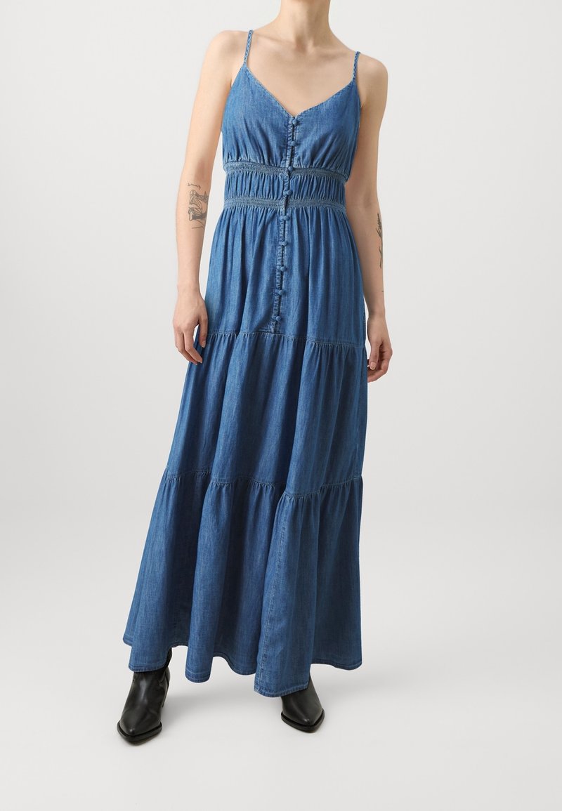 Personne portant une robe longue en jean bleu à fines bretelles et des bottes noires, debout devant un fond clair uni. Tatouages visibles sur les deux bras.