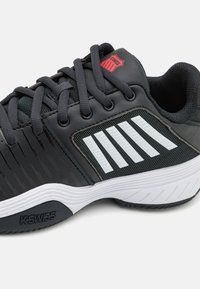 Zapato deportivo negro con acentos grises, que presenta materiales de malla y sintéticos, rayas texturizadas y una suela de goma con el logo de K-Swiss.