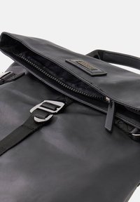 Schwarzer Rucksack mit glatter Textur, Reißverschlussöffnung und verstellbaren Trägern. Enthält eine silberne Schnalle und ein Markenlabel auf der Klappe.