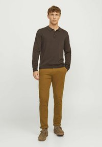 Polo marron à manches longues avec trois boutons, associé à un pantalon chino slim jaune moutarde et des chaussures en daim marron, se tenant devant un fond blanc.
