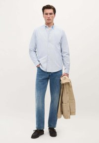 Camisa de botones a rayas azul claro, jeans de denim de corte relajado y una chaqueta beige colgada de un brazo. Zapatos negros completan el atuendo.