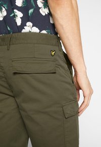 Homme portant un short cargo vert olive avec des poches à rabat et une chemise fleurie, main reposant près de la poche latérale.