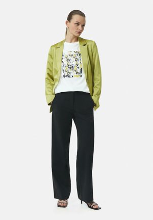 Groene satijnen blazer over een witte graphic t-shirt met bloemdesign, zwarte wijde broek en zwarte sandaaltjes met bandjes. Zachte texturen en een getailleerde pasvorm.