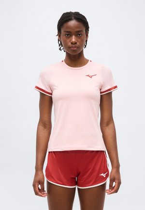 Nő világos rózsaszín rövid ujjú sportfelsőben, piros szegéllyel, valamint hozzáillő piros sportnadrág fehér szegéllyel, egy egyszerű háttér előtt áll.