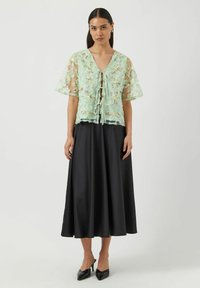 Mintgrønn gjennomsiktig bluse med blomsterbroderi, knyting foran, og korte ermer, kombinert med et svart satengskjørt og spisse svarte hæler.