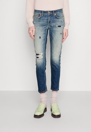 Frau trägt hellrosa Pullover, ausgewaschene blaue Jeans mit Used-Effekten, rosa Socken und blassgrüne Slipper, steht vor weißem Hintergrund.