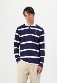Polo à manches longues bleu marine avec des rayures blanches horizontales. Il arbore un col blanc et des détails rouges avec le texte "HACKETT 1."