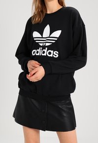 Svart sweatshirt med en vit Adidas-logotyp, med rund halsringning och ribbade ärmslut. Kombinerad med en svart läder minikjol.