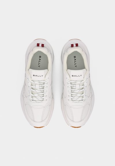 Paire de baskets blanches Bally avec lacets, étiquettes de languette brandées, languettes rouges au talon, et semelles légèrement texturées, vues de dessus sur un fond gris.