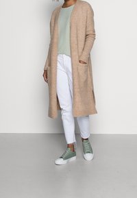 Cardigan long beige, texture côtelée, devant ouvert, porté sur un haut vert clair et un jean blanc, assorti à des baskets vert menthe.