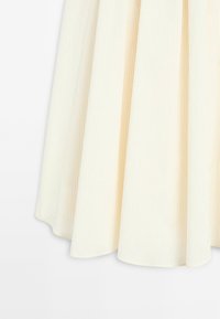 Jupe plissée de couleur crème clair avec des rayures verticales discrètes, fabriquée en tissu doux. Présente une silhouette fluide avec un ourlet qui s'incurve doucement.