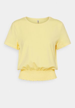 Keltainen lyhythihainen lyhyt paita, jossa on joustava, rypytetty vyötärönauha ja pyöreä pääntie, yksivärisellä valkoisella taustalla.