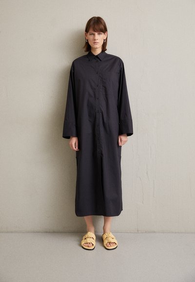 Schwarzes Langarmhemdkleid mit Knöpfen und einer Brusttasche, kombiniert mit gelben, geknoteten Slipper-Sandalen. Glatte Stoffoberfläche.