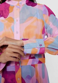 Blusa trasparente con motivi astratti nei colori rosa, arancione e blu. Presenta un colletto alla mandarino, bottoni e maniche lunghe con polsini a contrasto.