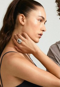 Anillo de plata con una superficie lisa y reflectante llevado en una mano, mostrando un perfil en primer plano de una persona contra un fondo plano.