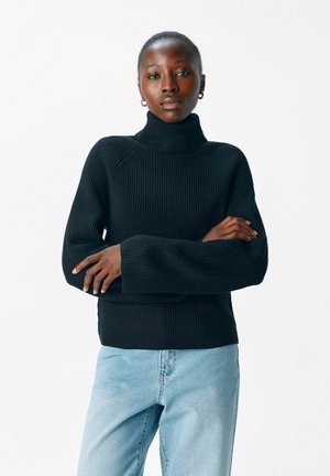 Pull à col roulé noir côtelé avec des manches allongées, présentant un design ajusté et un tricot texturé. Associé à un jean bleu clair.