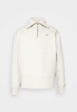 Beige vetoketjullinen collegepaita, jossa on etutasku, yhdistetty sinivalkoisiin abstraktikuviollisiin housuihin. Asun täydentävät mustat slip-on-kengät.