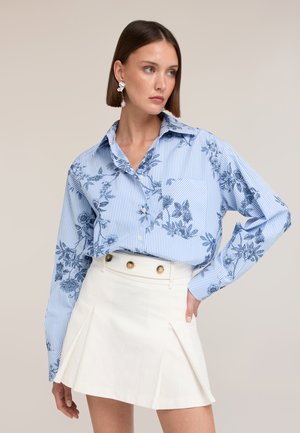 Femme aux cheveux courts et bruns portant une chemise rayée bleue et blanche à motif floral rentrée dans une jupe plissée blanche taille haute avec des boutons dorés.