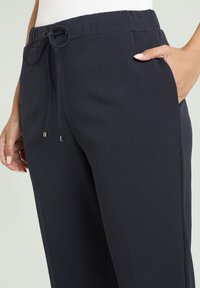 Detailní záběr osoby v tmavě modrých kalhotách na šňůrku, s jednou rukou v kapse na světlém pozadí.