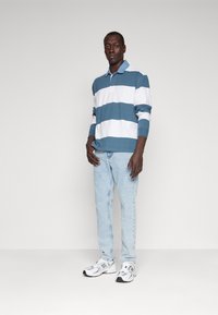 Bărbat în picioare, îmbrăcat cu un polo cu dungi albastre și albe cu mânecă lungă, blugi ușor albaștri și adidași New Balance albi, pe un fundal simplu.
