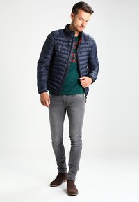 Mørkeblå puffjakke med høj krave, båret over en grøn skjorte, parret med grå slim-fit jeans og brune ankelstøvler. Glat tekstur og quiltet design.