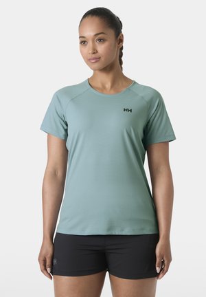 Młoda kobieta w jasnorozowym sportowym t-shircie z krótkim rękawem i czarnych szortach, stojąca na tle jednolitego białego tła.