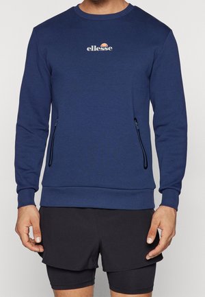 Mężczyzna w niebieskiej bluzie ellesse z kieszeniami na zamek i czarnych sportowych szortach na jednolitym tle.