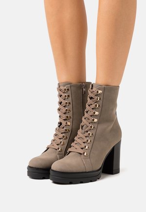 Botas de tobillo de imitación cuero marrón con un alto tacón cuadrado, que presentan ojales dorados, cordones y un zipper lateral. Acabado texturizado y cuello acolchado.