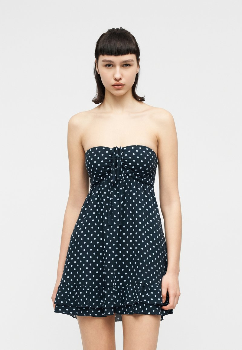 Νεαρή γυναίκα με κοντά σκούρα μαλλιά, που φοράει μια μπλε ναυτικό φόρεμα χωρίς τιράντες με λευκές πουά και βολάν στο τελείωμα, στέκεται μπροστά σε απλό φόντο.