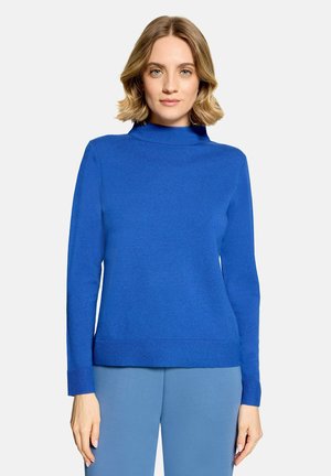 Femme aux cheveux blonds courts portant un pull bleu vif à manches longues et un pantalon bleu clair, debout devant un fond uni clair.