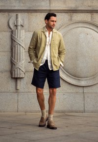 Homme portant une veste beige, une chemise blanche, un short bleu marine et des chaussettes beiges avec des mocassins, debout contre un mur en pierre orné de sculptures décoratives.