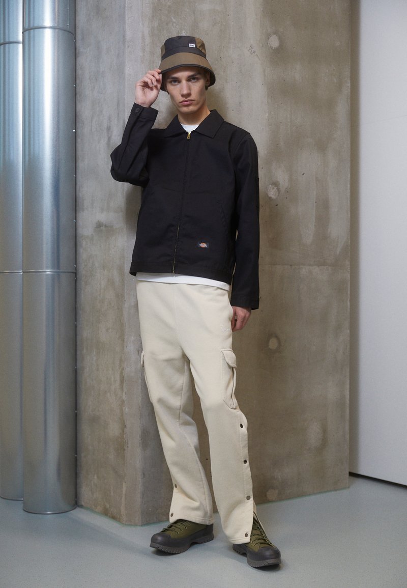 Giacca leggera nera con zip sul davanti, pantaloni cargo beige con tasche laterali e chiusure a scatto laterali, scarponi da trekking verdi e un cappello a secchiello marrone.
