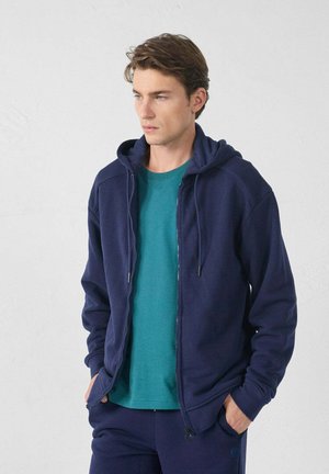 Donkerblauwe zip-up hoodie met een trekkoordcapuchon, gecombineerd met een teal ronde hals t-shirt. Gladde stof met geribbelde accenten op de mouwen.