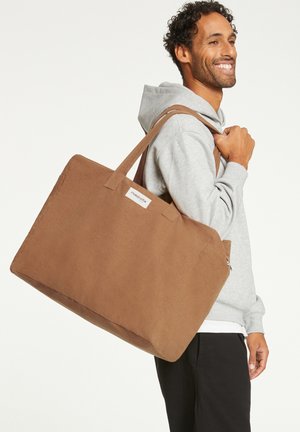 Brown tote bag with a soft texture, rectangular shape, dual handles, and a label on the side. Suitably sized for everyday use.