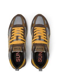 Baskets marron et grises avec des lacets jaunes, en tissu et cuir, de forme arrondie, logo sur la languette et détails texturés sur les côtés.
