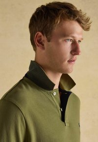 Olivegrünes Polohemd mit einem gestreiften schwarzen Kragen, ausgestattet mit einer Knopfleiste mit zwei Knöpfen und einem dezenten Logo auf der Brust, strukturiertem Stoff.