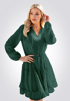 Abito verde a strati con maniche lunghe, scollo a V e coulisse. Presenta pois testurizzati e orlo arricciato. Tessuto setoso. Collana in catena dorata.