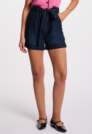 Dark denim shorts with a high waist, rolled cuffs, and a fabric tie belt. Paired with a pink top and black pointed flats.