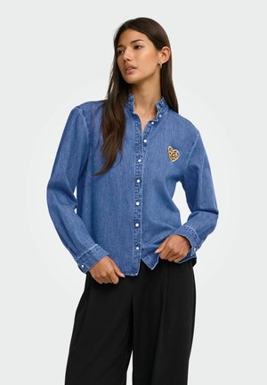 Femme aux longs cheveux foncés portant une chemise en denim bleue boutonnée avec un col à volants et un patch en forme de cœur imprimé léopard, associée à un pantalon noir.