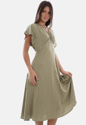 Donna con capelli lunghi e scuri che indossa un vestito midi verde oliva chiaro con maniche corte e un delicato motivo a pois, tenendo il lato della gonna.