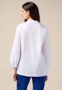 Blusa blanca de manga larga con bordado texturizado y un ajuste relajado, que presenta un cuello alto y un ligero acampanado en el dobladillo.