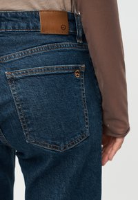 Πίσω όψη ενός ατόμου που φοράει σκούρο μπλε τζιν τζιν και καφέ μακρυμάνικη μπλούζα, εμφανίζοντας τις λεπτομέρειες της πίσω τσέπης και της μέσης.