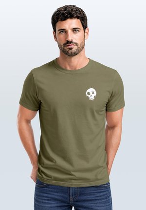 Mann mit kurzen lockigen Haaren, der ein olivgrünes T-Shirt mit einer kleinen weißen tropfenden Totenkopf-Grafik auf der linken Brust trägt, steht mit den Händen hinter dem Rücken.