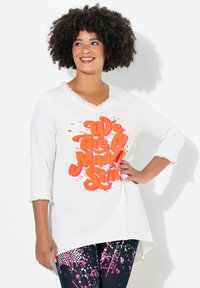 Witte langemouwen shirt met een versleten halslijn en heldere oranje tekst die "We Are All Mad Here" leest. Draagt met een patroon van donkere legging.