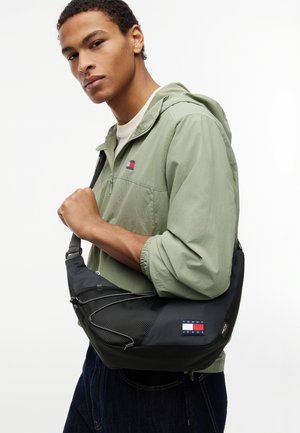Młody mężczyzna w jasnozielonej kurtce z kapturem trzyma na ramieniu czarną torbę na ramię Tommy Jeans na jednolitym tle.