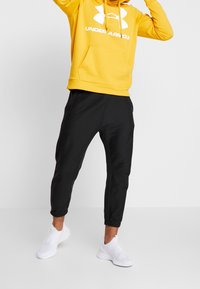 Sudadera amarilla con capucha y gran logo blanco, combinada con joggers negros ajustados y zapatillas deportivas blancas sin cordones.
